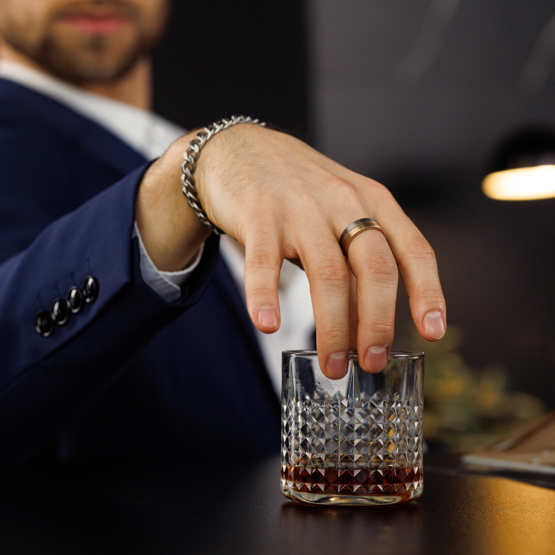 Man i blå kostym lyfter whiskeyglas, ringen i roséguld blänker i barbelysning.