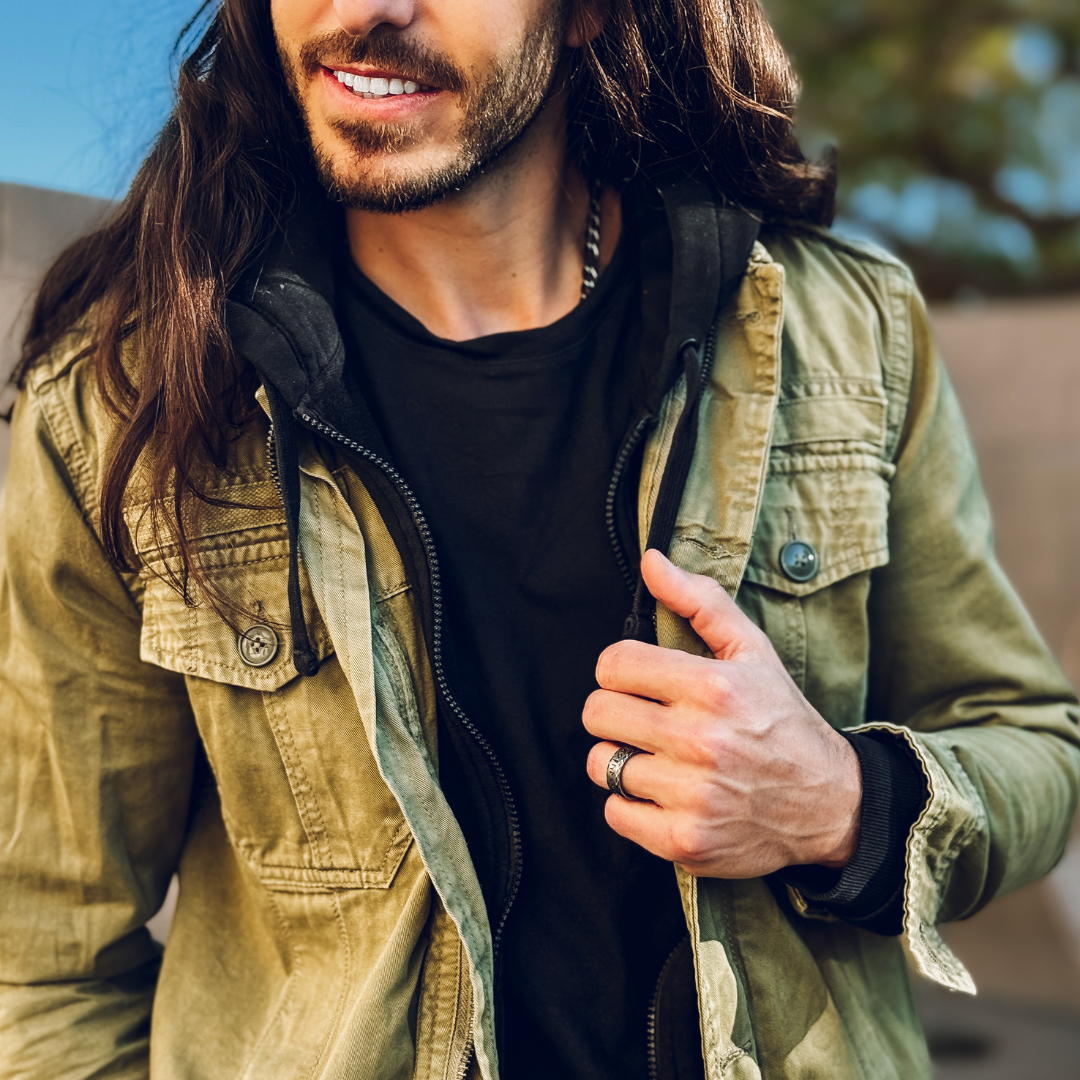 Långhårig man med rustik stil bär ring med runor, blickar ned i avslappnad pose.