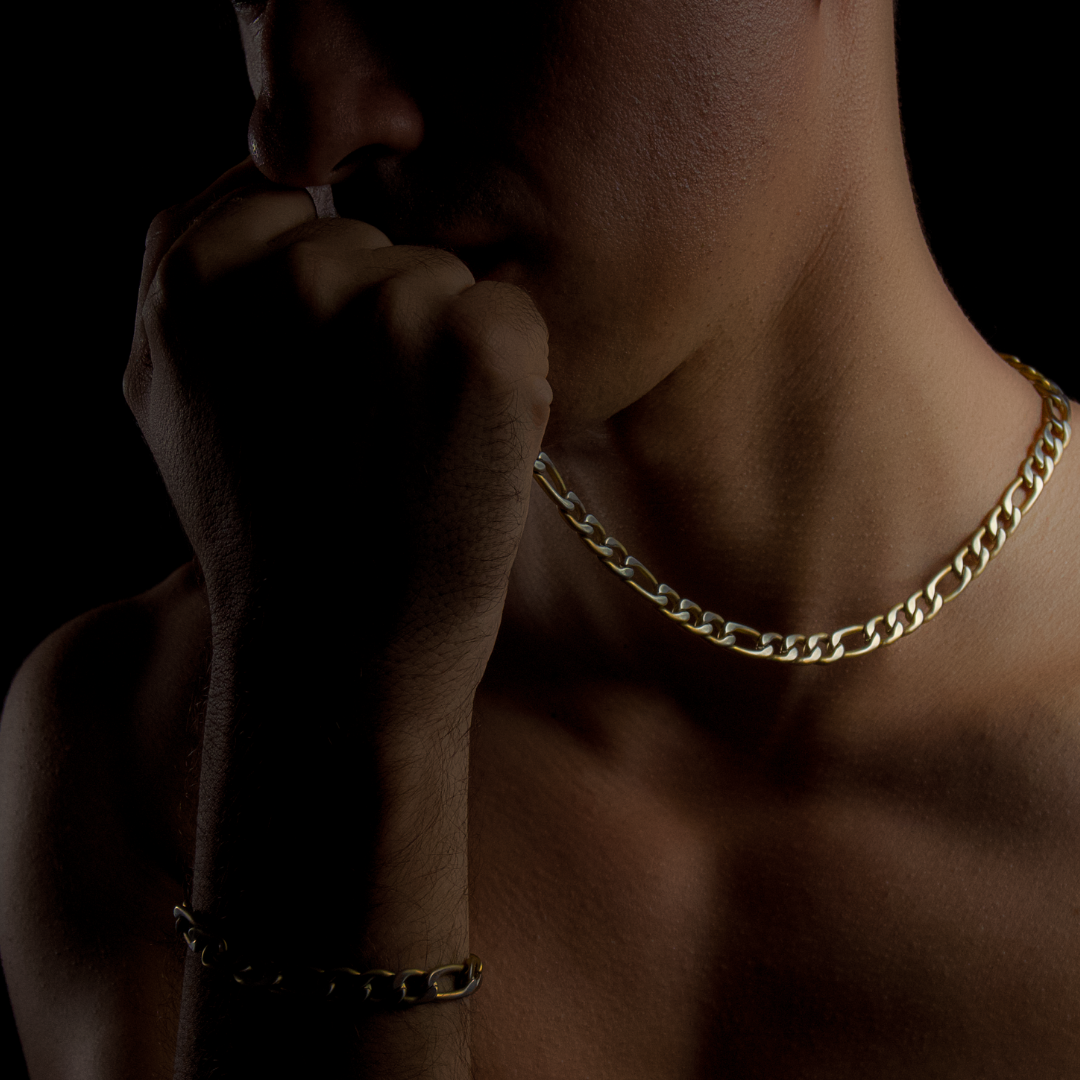 Kedjan i fokus på bar bröstkorg, mjukt ljus framhäver länkarnas form.#en:color_gold,sv:farg_guld