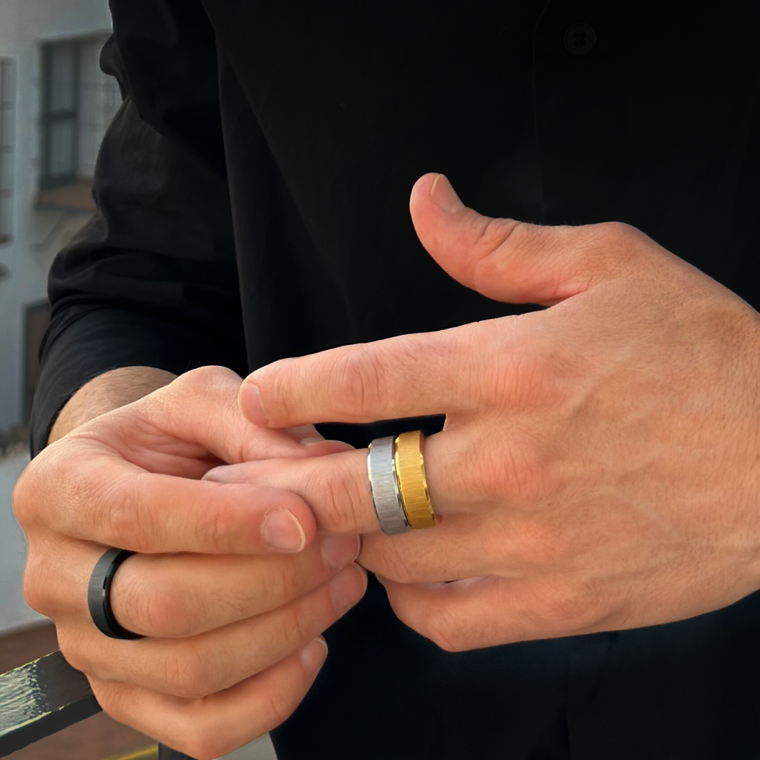 Närbild på händer som trär på en stilren silver- svart- och guldfärgadring, svart skjorta i bakgrunden.#en:color_silver,sv:farg_silver