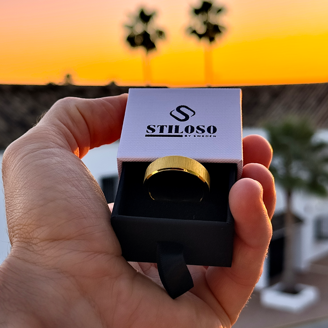 Hand håller upp en ringask med guldfärgad stålring framför solnedgång.#en:color_gold,sv:farg_guld