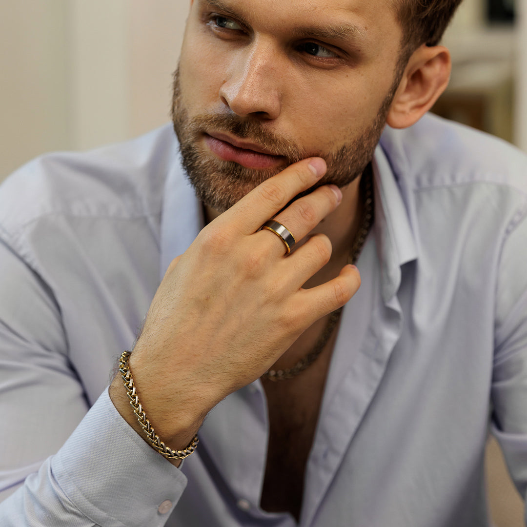 Man wearing a light blue shirt with a gold bracelet and ring, looking thoughtful.
#en:color_silver,sv:farg_silver