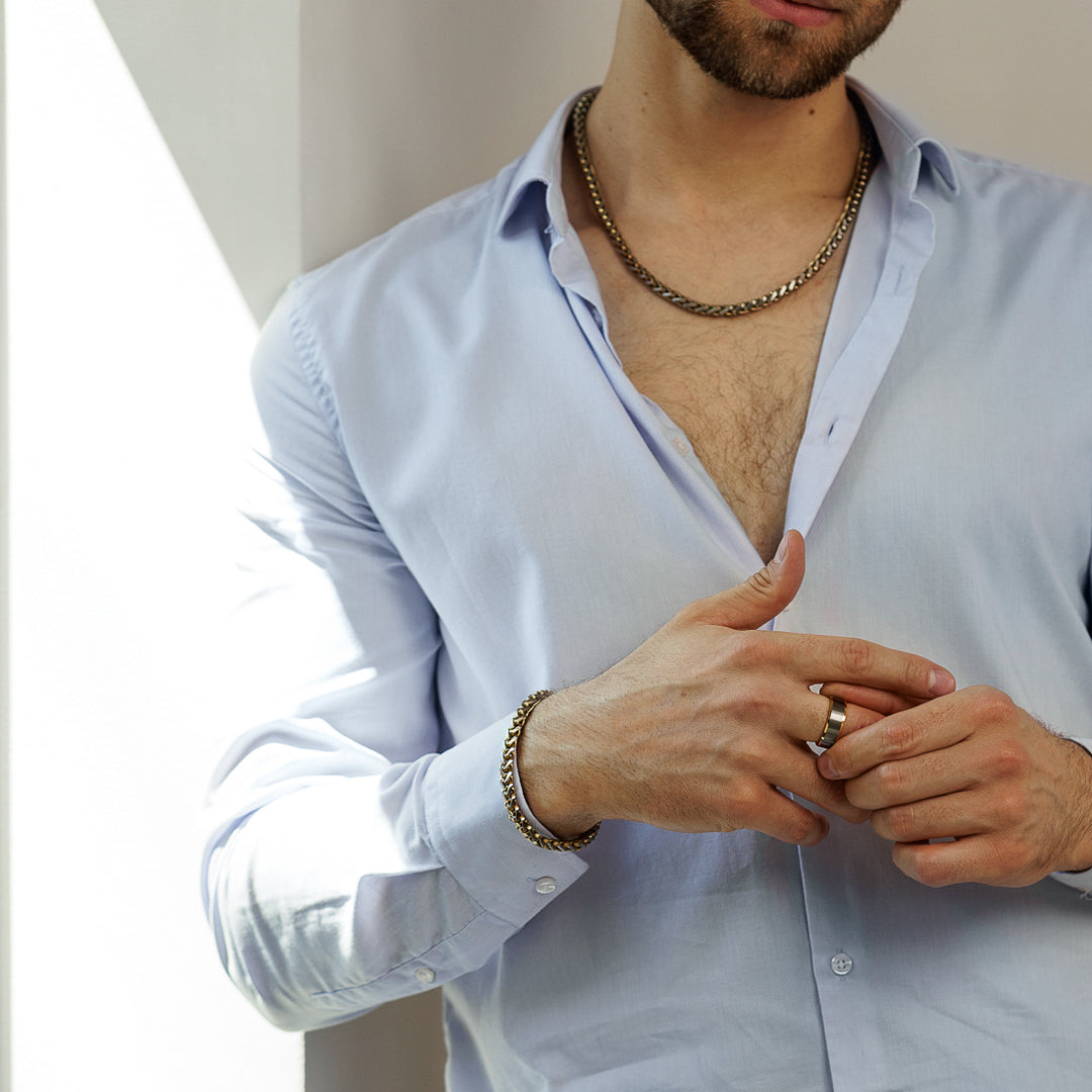 Man wearing a light blue shirt with rolled-up sleeves, gold chain necklace, and bracelet.
#en:color_gold,sv:farg_guld