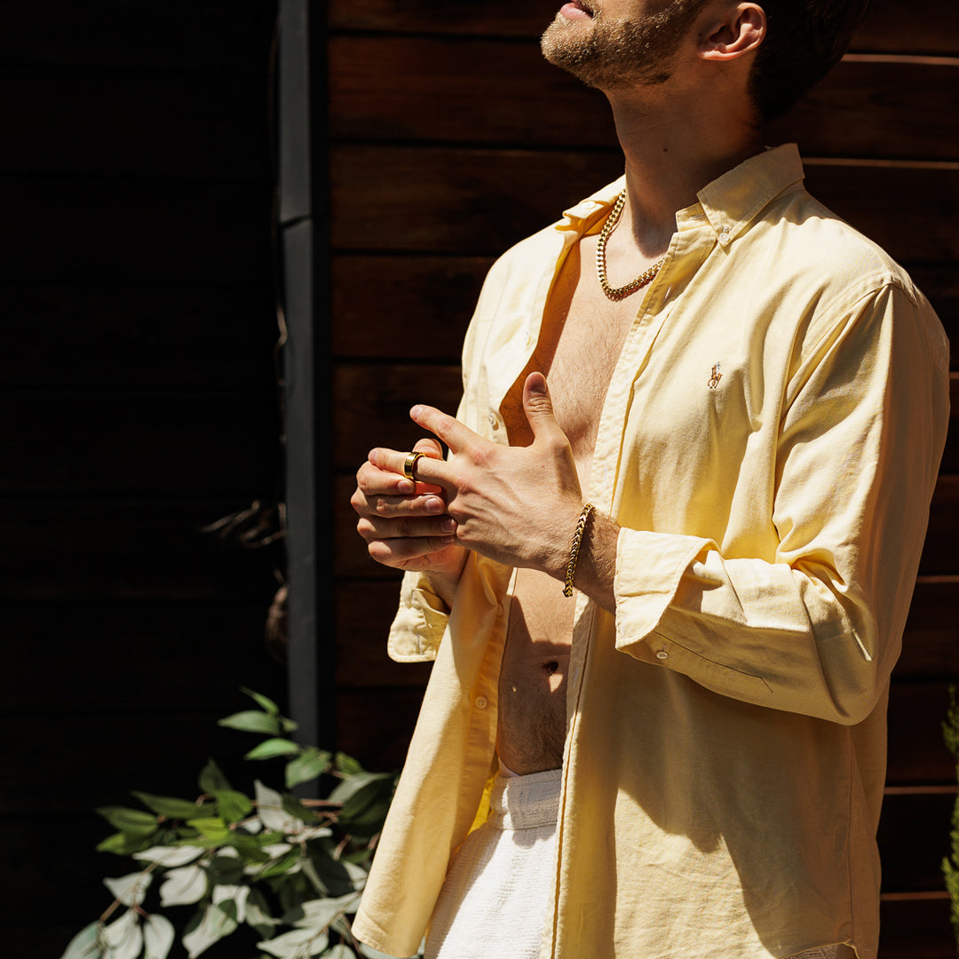 Man wearing a yellow shirt and white pants standing outdoors with a wooden wall and plants in the background.
#en:color_gold,sv:farg_guld