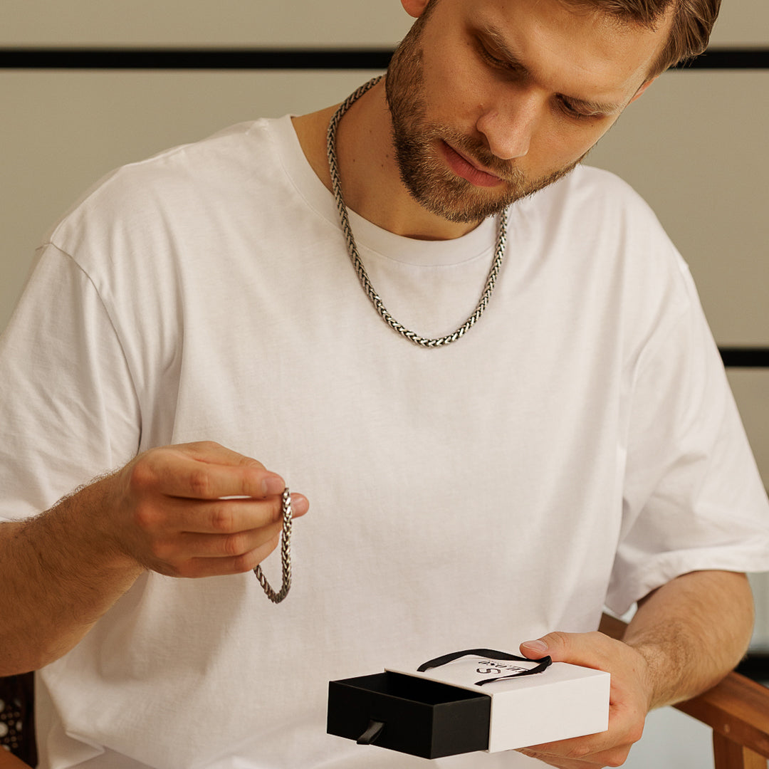 Man holding a silver chain and a jewelry box with a neutral background.
#en:color_silver,sv:farg_silver