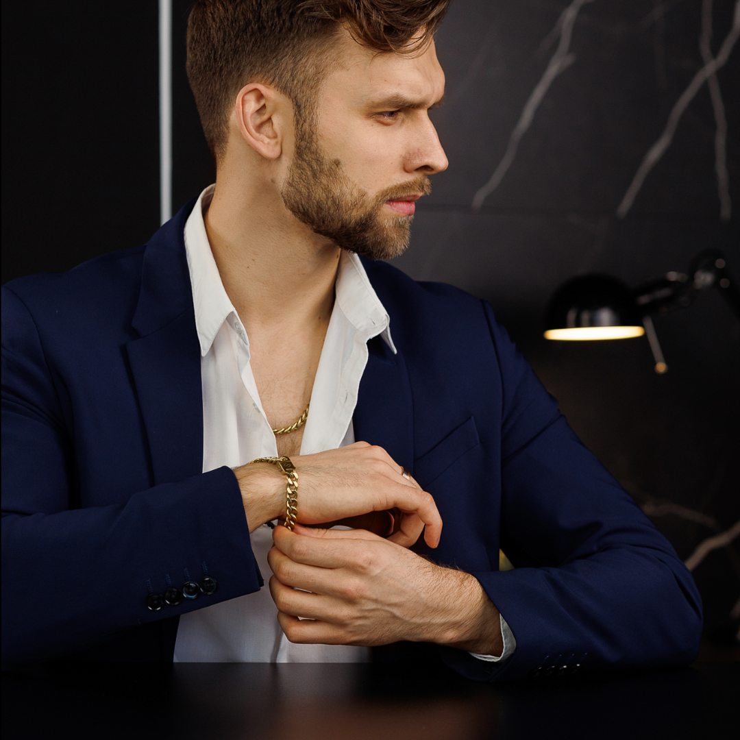 Man i marinblå kostym bär guldfärgat cuban link-armband och kedja, elegant look i mörk miljö.#en:color_gold,sv:farg_guld