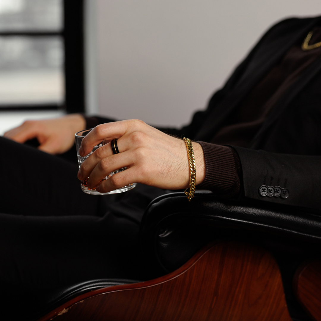 Man vilar handen på armstödet, guldfärgat armband syns mot mörk tröja.#en:color_gold,sv:farg_guld