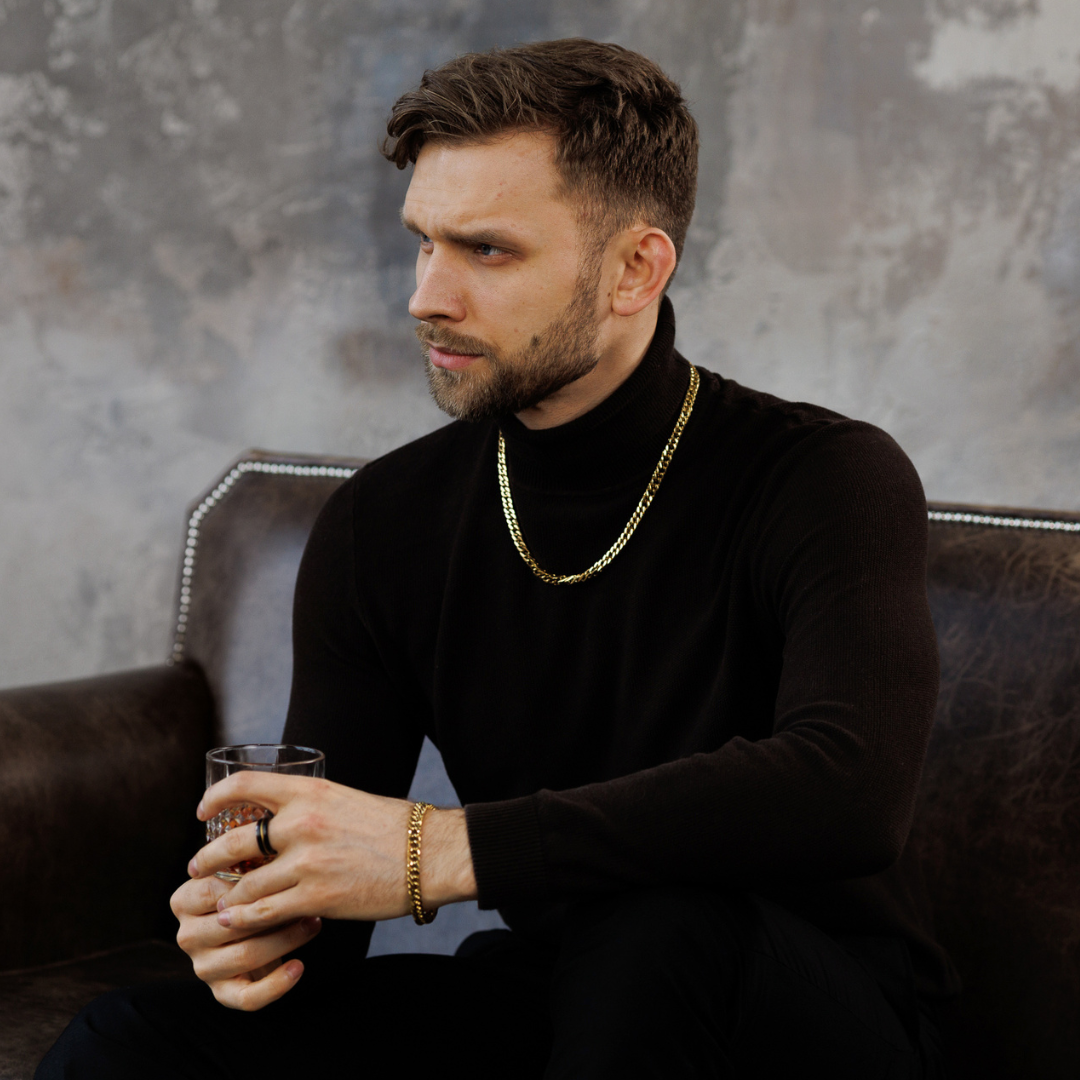 Man i svart tröja med guldsetet, sitter med whiskyglas i handen – elegant och självsäker vibe.#en:color_gold,sv:farg_guld