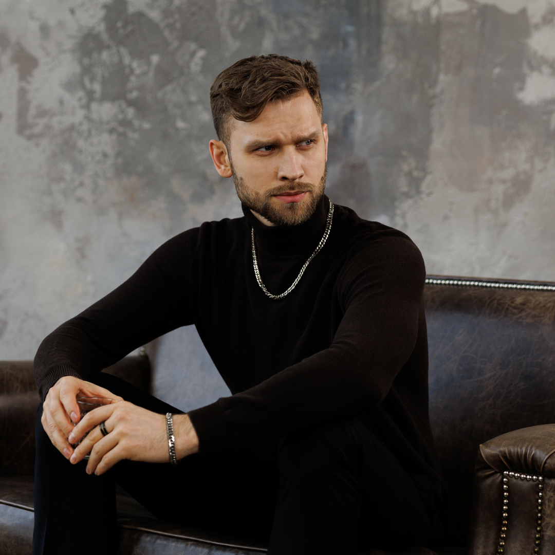 Sittande man i mörk klädsel, silverkedjan och armbandet adderar modern skärpa.#en:color_silver,sv:farg_silver