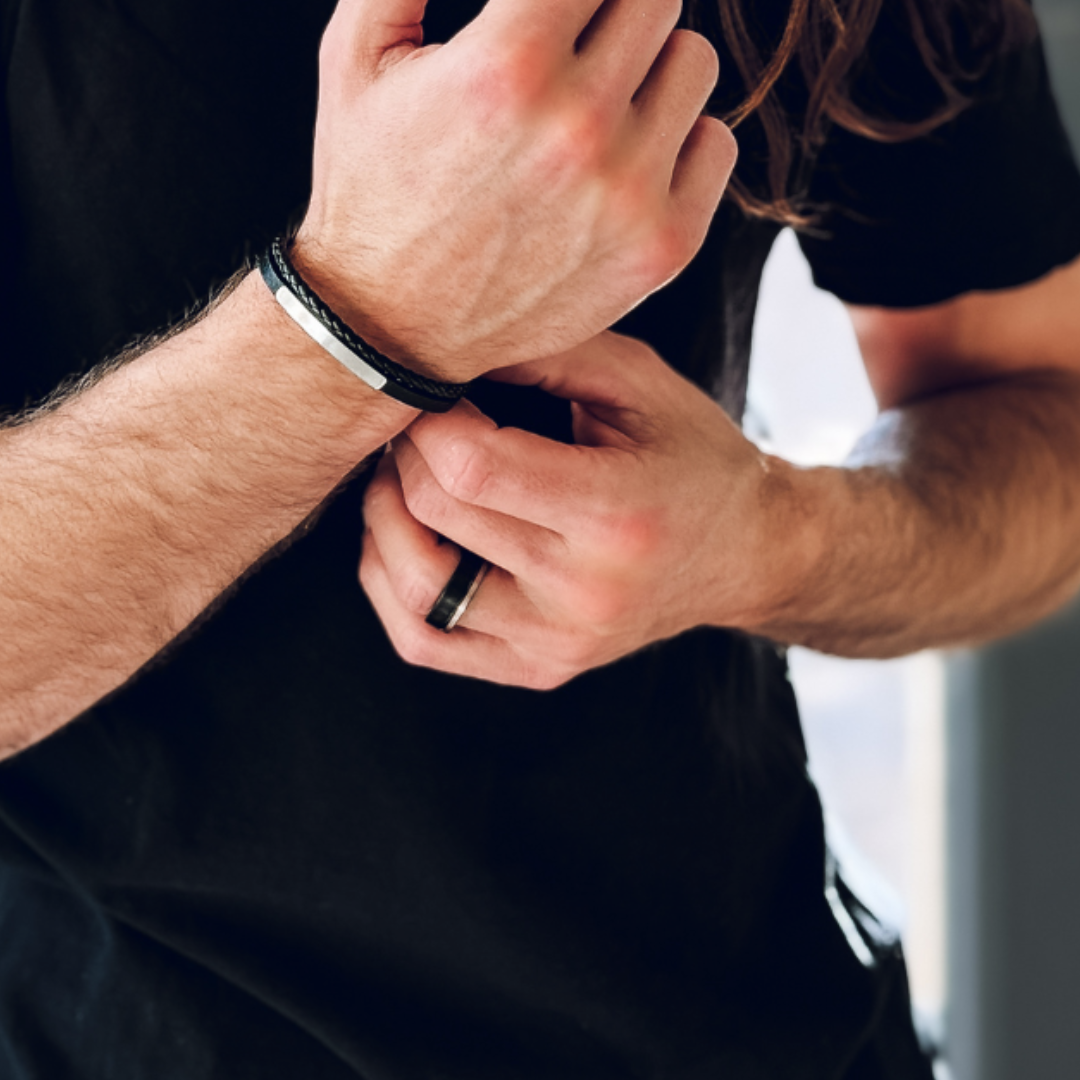 Man i svart t-shirt bär mörk ring och armband, händerna justeras i en vardaglig gest.