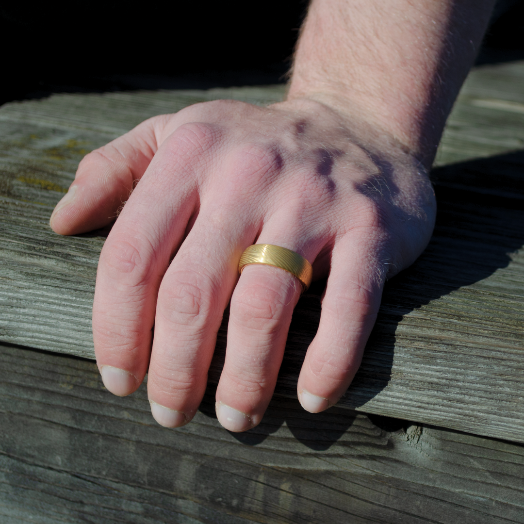 Ringen glänser i dagsljus när handen vilar mot träyta utomhus.#en:color_gold,sv:farg_guld