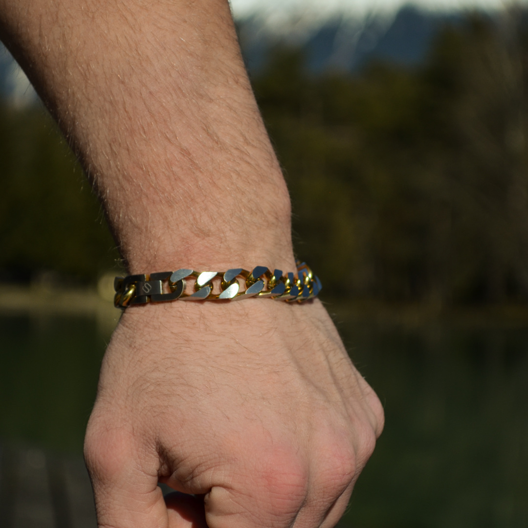 Armbandet glänser i solljus mot bar handled, utomhusmiljö med naturlig bakgrund.#en:color_gold,sv:farg_guld