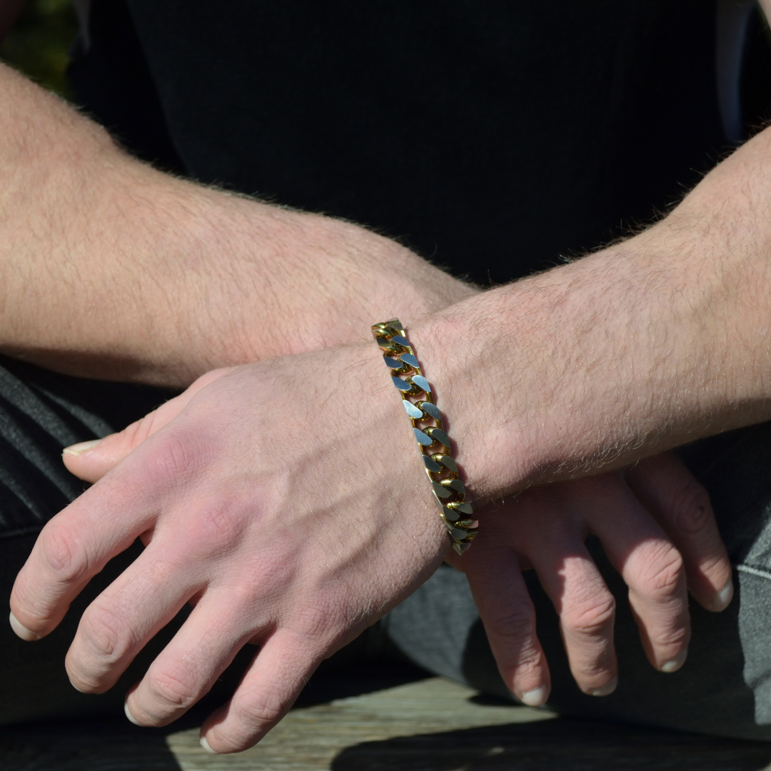 Armbandet i guld och silver reflekterar solljus, vilande över korslagda handleder i avslappnad pose.#en:color_gold,sv:farg_guld