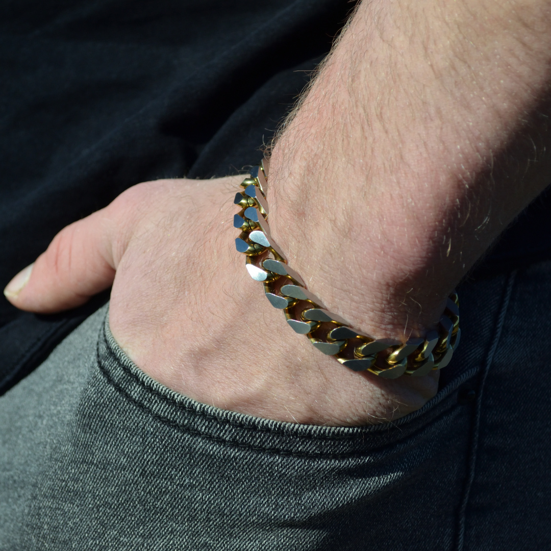 Närbild på handled med guldfärgat cuban link-armband, handen i fickan mot jeans och svart t-shirt.#en:color_gold,sv:farg_guld