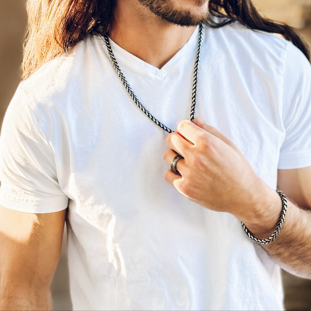 Avslappnad man bär svart- silverarmband och kedja, fångad i mjukt solljus och vit t-shirt.#en:color_black,sv:farg_svart