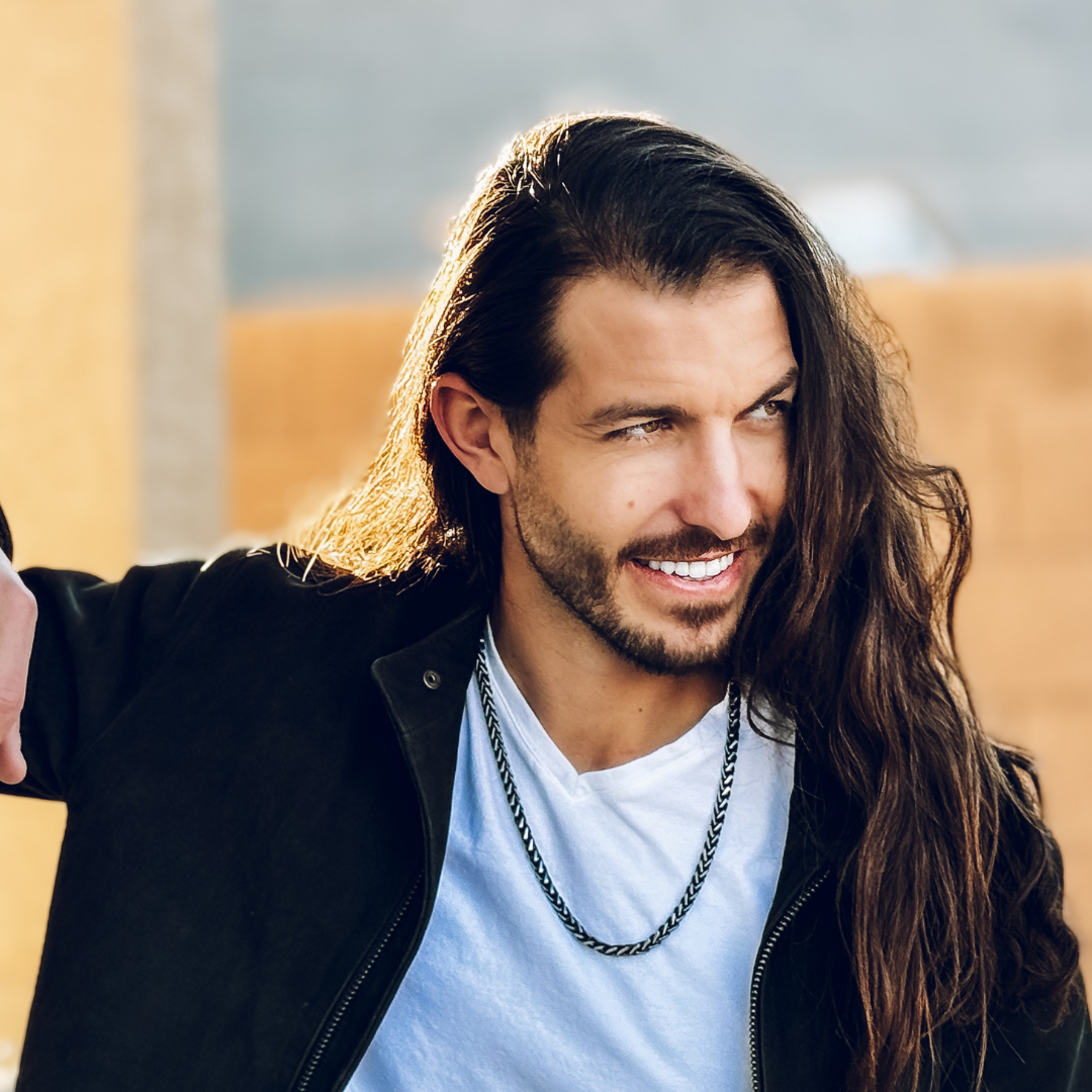 Långhårig man i svart jacka och vit tröja bär kedjan med självsäker blick.
#en:color_black,sv:farg_svart